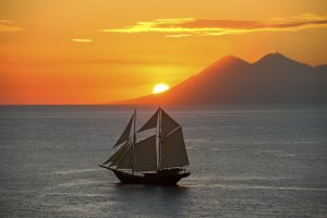 luxusschiff amandira auf hoher see mit gesetzten segeln bei sonnenuntergang vor einem berg der auf dem festland vor der untergehenden sonne zu erblicken ist