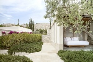 Blick auf die Veranda eines Pavilion Bedrooms im Amanzoe in einer wunderschönen Gartenanlage und hellem Stein rechts ist das Bett des Pavilion Zimmers zu sehen Luxusresort Griechenland