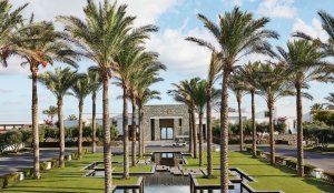 eingang des luxushotels amirandes kreta mit vielen palmen und flachem brunnenverlauf führt direkt zum eingang der mit naturstein inmitten des garten liegt