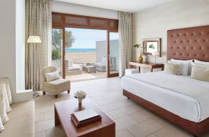 helles schlafzimmer einer strandvilla im hotel amirandes mit warmen farbtönen und blick auf den strand und das blaue meer durch die großen fenster die zur terrasse führen
