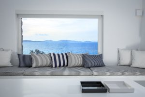gemütliche sitzecke der lounge in der privaten villa big blue beach villa auf mykonos mit direktem blick auf das meer durch das große fenster hinter der loungeecke