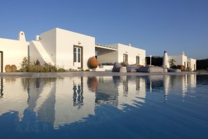 blick über den großen pool der privaten villa erosantorini und die villa mit ihren weißen bauten unter strahlend blauem himmel direkt am meer