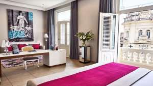 geräumige junior suite mit modernen und hellen möbeln und pink farbene elemente mit vielen fenstern neben dem bett und dem wohnbereich