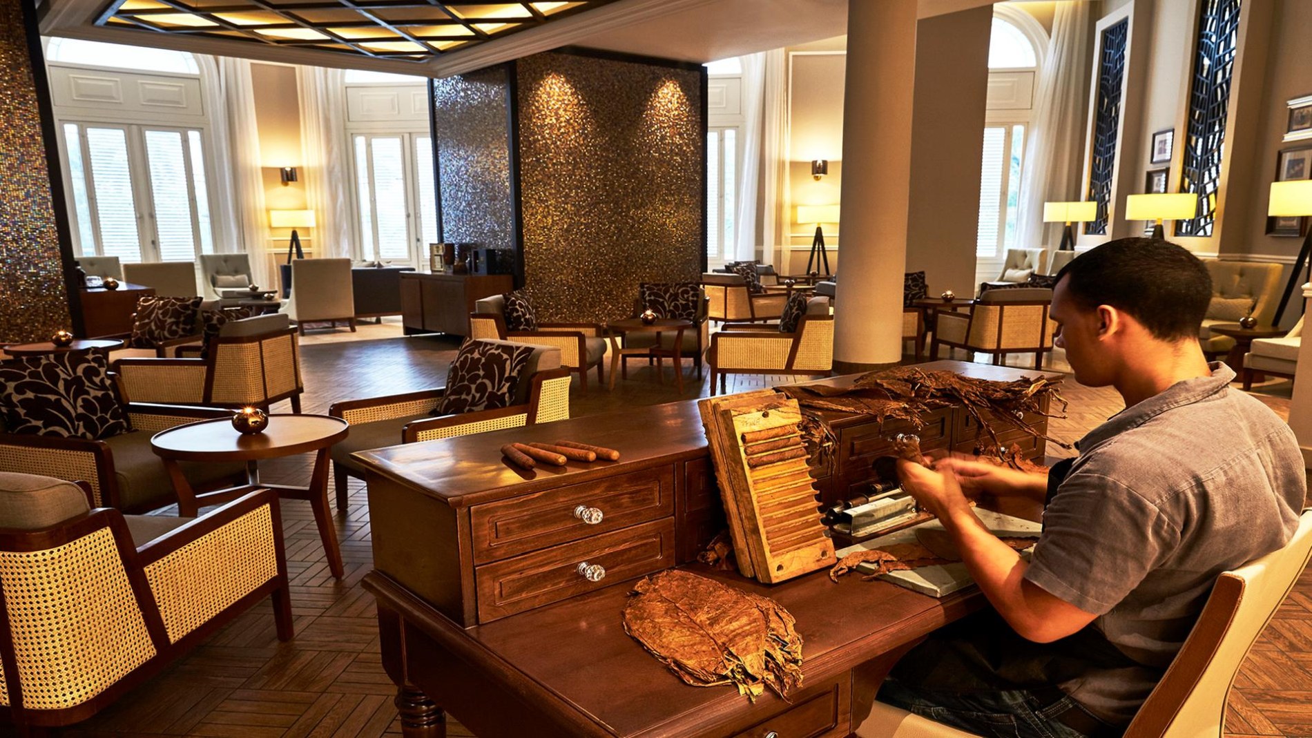 gemütliche zigarren lounge mit dunklen hölzern und beigen stoffen eingerichtet mit dem cigar desk im vordergrund an dem ein kubaner handgefertigte zigarren herstellt