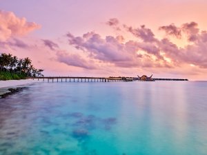 aussicht vom strand auf das joali malediven luxusresort direkt über dem wasser errichtet bei herrlichem sonnenuntergang im türkisfarbenem wasser und lila erstrahltem himmel