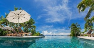 Blick über den großen azurblauen Luxus Pool des Likuliku Lagoon Resort mit saftig grünen Palmen umgeben unter strahlend blauem Himmel