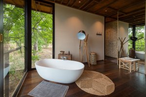 freistehende badewanne im luxushotel in botdswana direkt vor den großen fenstern mit blick in die landschaft von botswana mit warmen farbtönen im holzboden und den wänden