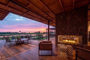 lounge und terrasse mit offenem kamin vor einer großen holzterrasse mit großem runden tisch und stühlen bei sonnenuntergang mit aussicht auf die natur von botswana und das wasser