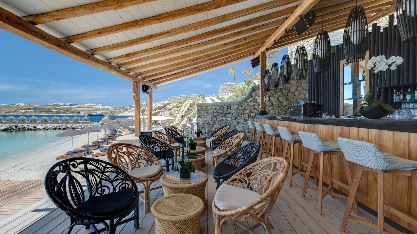 foto von einer überdachten strandbar mit stühlen und kleinen tischen alles aus holz am meer mit blick auf das kristall klare smaragd grüne wasser im luxuriösen hochklassigen exklusiven designer hotel santa marina auf mykonos in griechenland europa