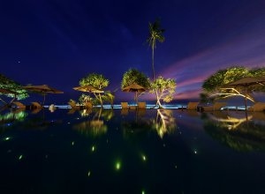 blick auf den strand von kogalla über den großen pool in dem sich lichter spiegeln im hintergund das blaue meer mit palmen und liegestühlen