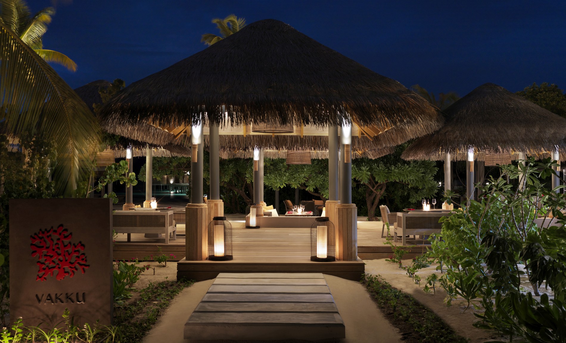 blick zwischen palmen auf die gemuehtlichen sitzmoebel an gedeckten tischen unter holzdaechern laden zum essen im luxus restaurant ein