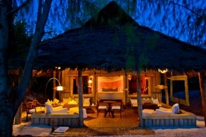 Außenansicht auf eine Bande bei Nacht mit Veranda und Bett inmitten des Raums mit romantischer Beleuchtung in der andBeyond Mnemba Island Lodge auf Sansibar 
