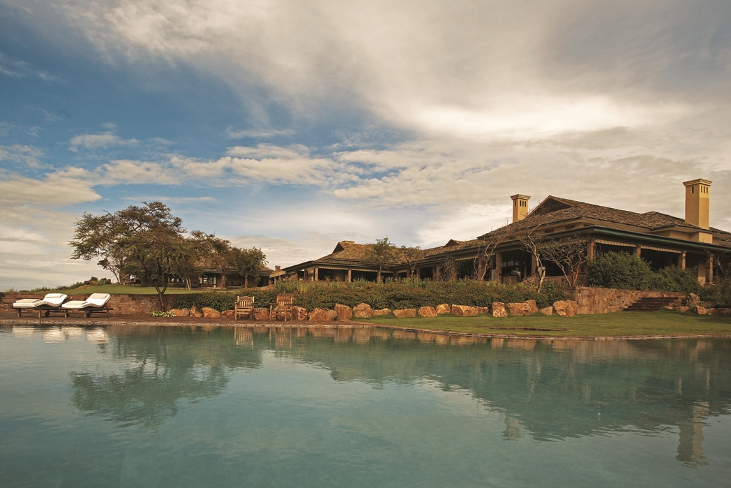 imposanter Pool der sasakwa lodge in afrika mit blauem wasser und den holzhäusern des luxushotels im hintergrund unter bewölktem himmel