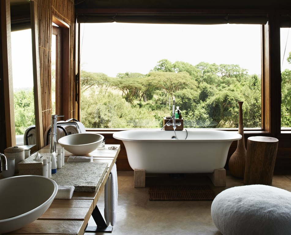 große freistehende badewanne vor einem rießigen fenster mit blick in die natur afrikas daneben zwei waschbecken mit hellem holz an der wand und als möbel